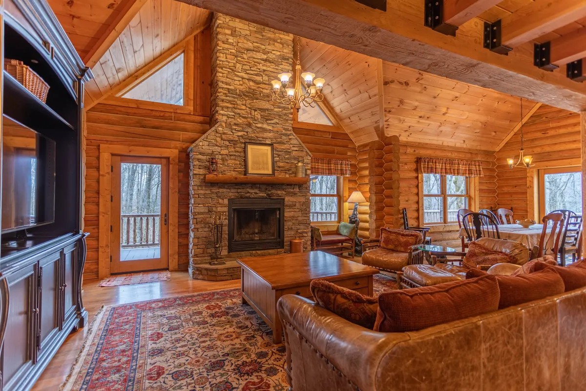 A different angle on the grand living area, this image highlights the stone fireplace, exposed beams, a dining table, and a wall of large windows flooding the space with natural light. The comfortable seating area invites relaxation, and the integration with adjacent dining spaces reinforces the idea of a social hub.