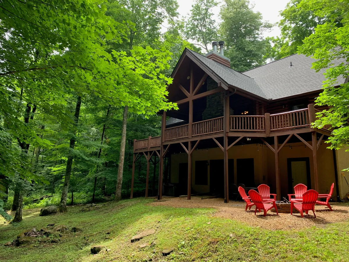 This exterior image features the log cabin structure set among surrounding trees, with red Adirondack chairs arranged invitingly in the foreground. The natural setting hints at mountain adventures and the cozy comforts of returning home after exploring outdoors.
