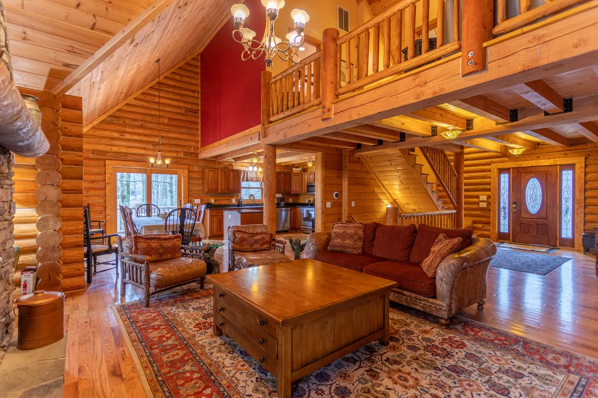 The view features a cozy, comfortable living area with a large wooden coffee table, soft sofas, a distinctive chandelier, and a partial glimpse into the kitchen area beyond. The staircase and open plan hint at easy movement and connection throughout the main living spaces, perfect for shared activities.