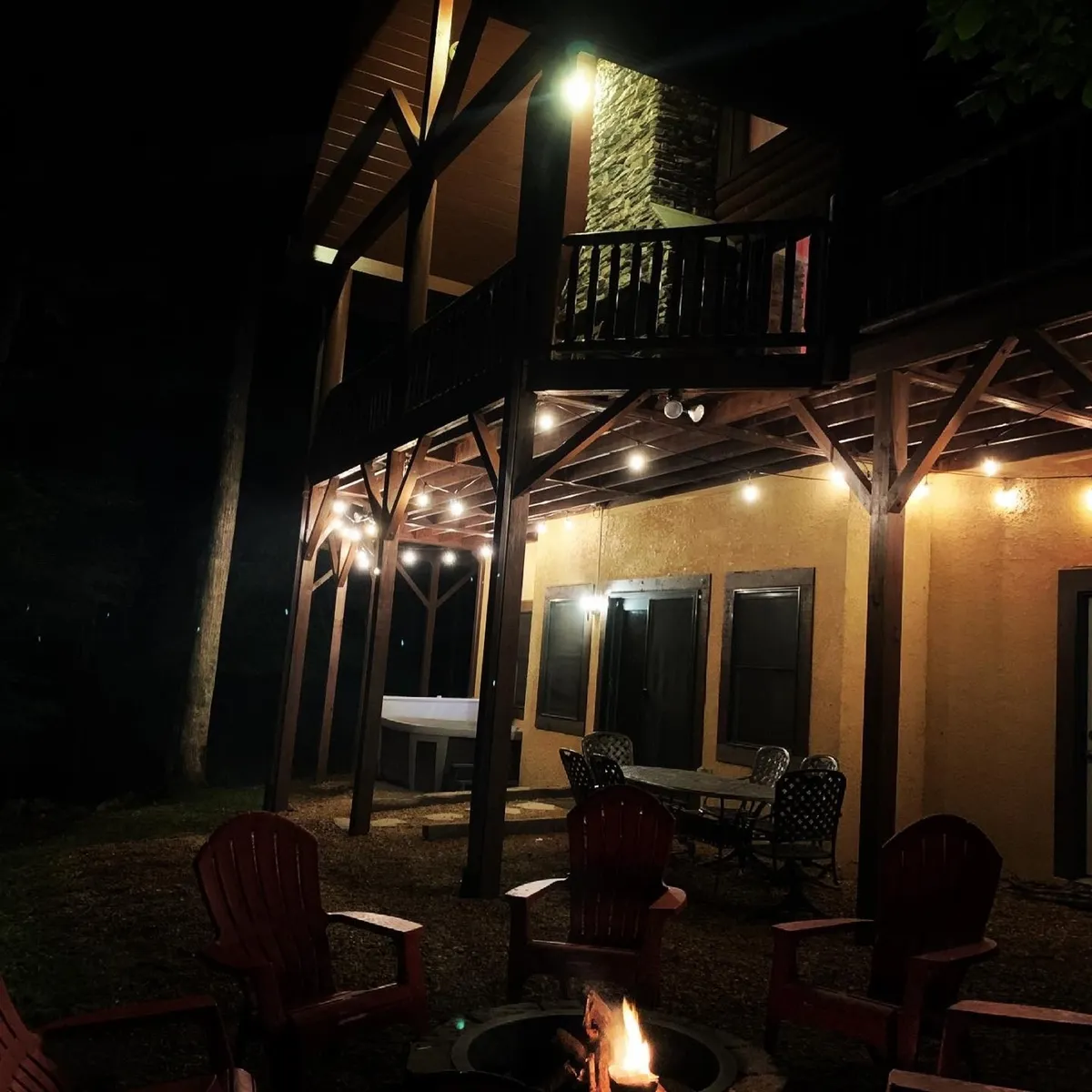 An inviting outdoor setting featuring a fire pit surrounded by Adirondack chairs, wooden beams strung with hanging lights, and a jacuzzi nestled in a private, wooded area. The scene is framed by lush greenery, creating a sense of seclusion and tranquility.