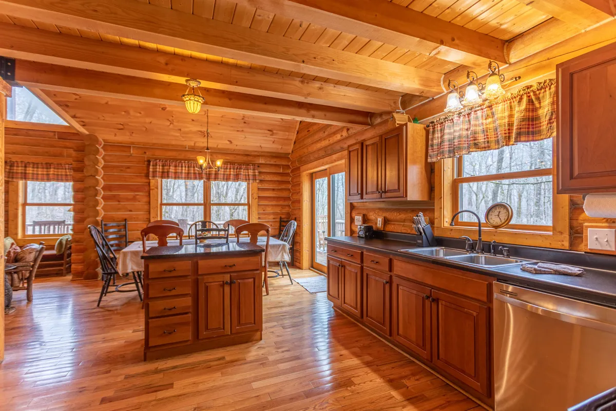 A large wooden dining table sits in an inviting space beside the kitchen, surrounded by log-cabin walls and illuminated by natural light pouring through expansive windows. The kitchen island is visible, reinforcing the connection between spaces.