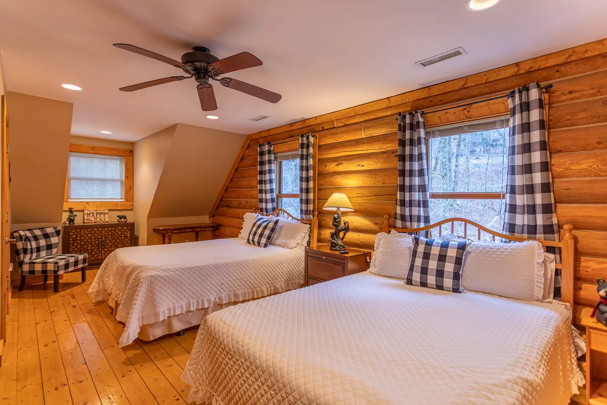 An inviting guest bedroom with two beds dressed in decorative pillows, wooden walls, ample natural light from windows with checkered curtains, and a bedside lamp for nighttime reading.