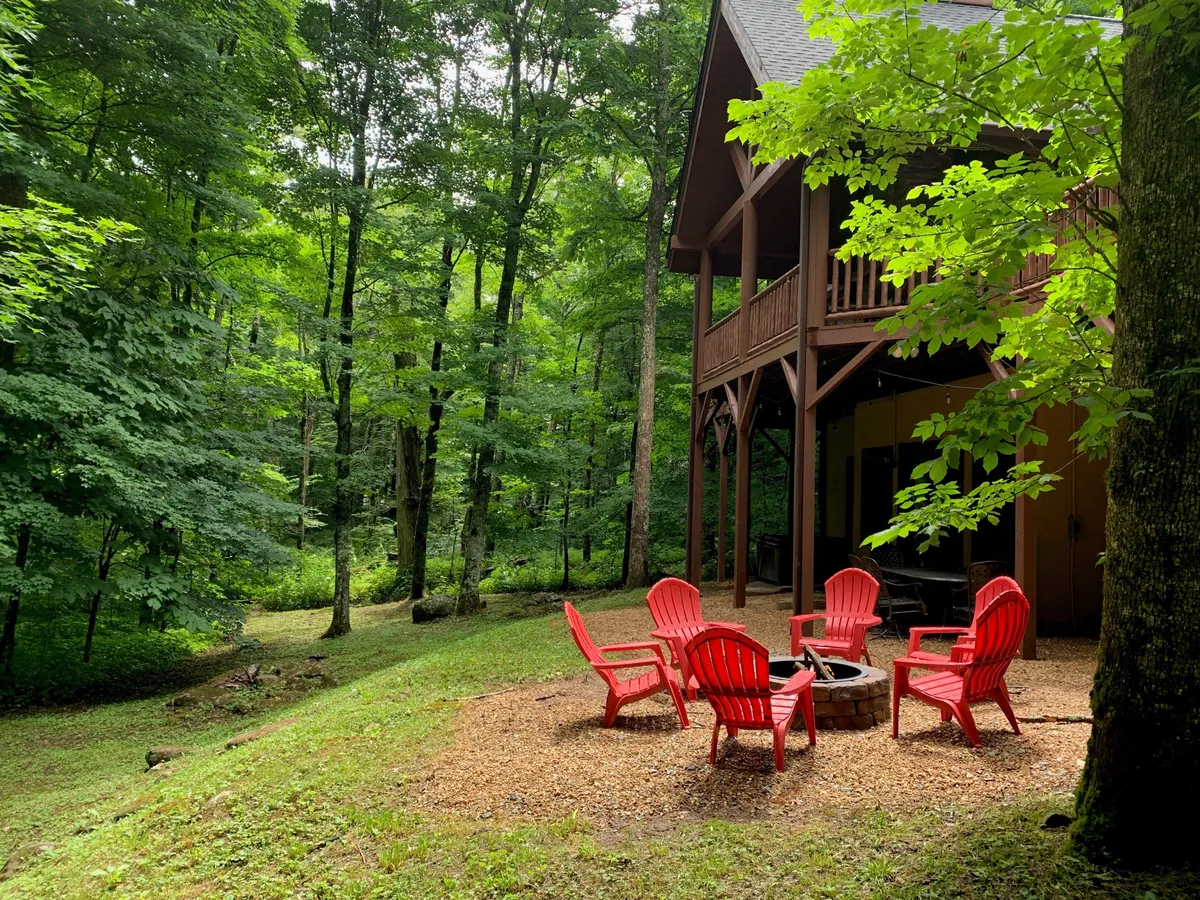 This image features a welcoming outdoor fire pit area framed by red Adirondack chairs, surrounded by mature trees and a rustic wooden structure. The setup is ideal for group gatherings, storytelling, and relaxing in nature, capturing the essence of communal fun and evening camaraderie.