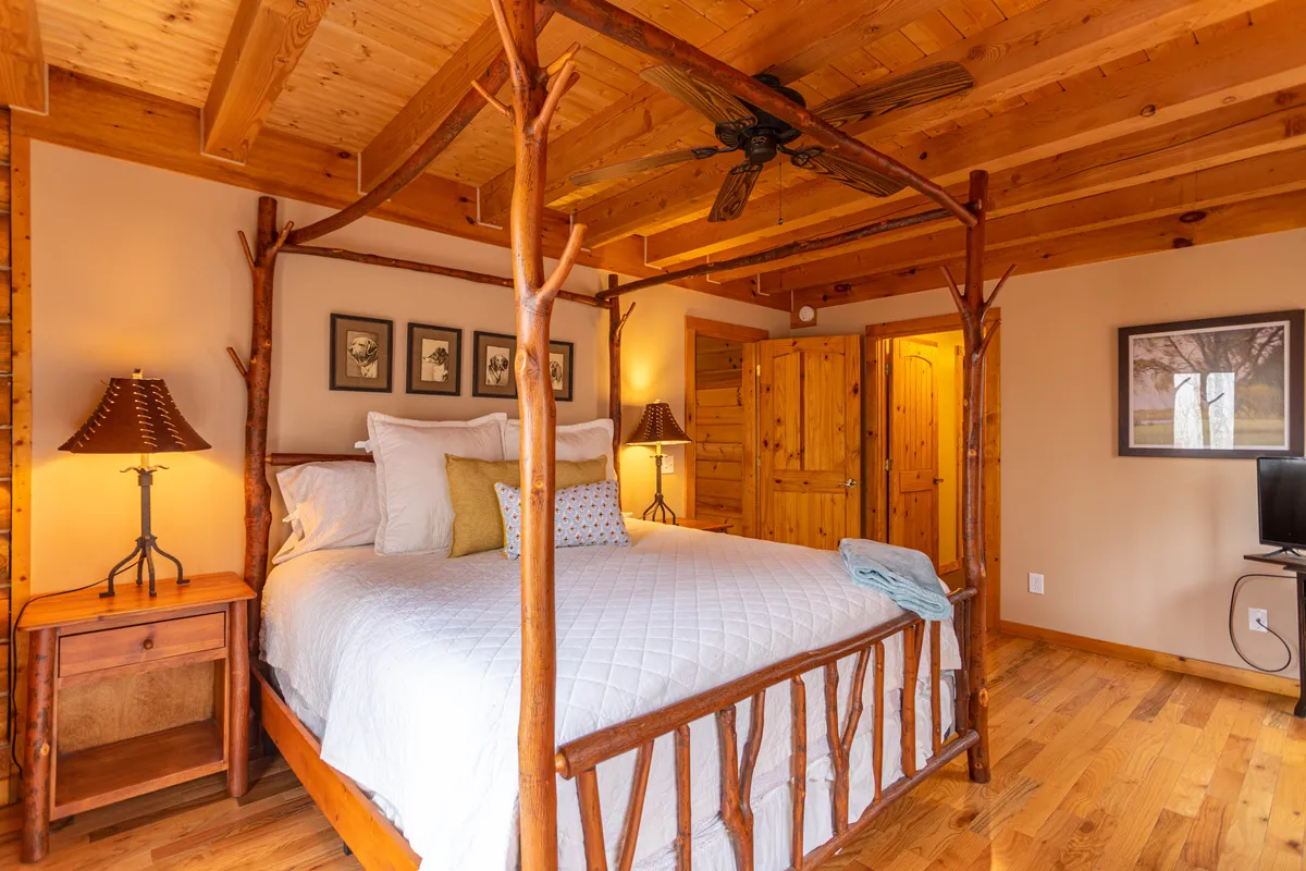 A serene master bedroom featuring a distinguished four-poster king bed, wooden ceiling beams, and soft lamps on bedside tables. Natural light fills the space, and the decor exudes rustic elegance, ideal for a restful retreat.