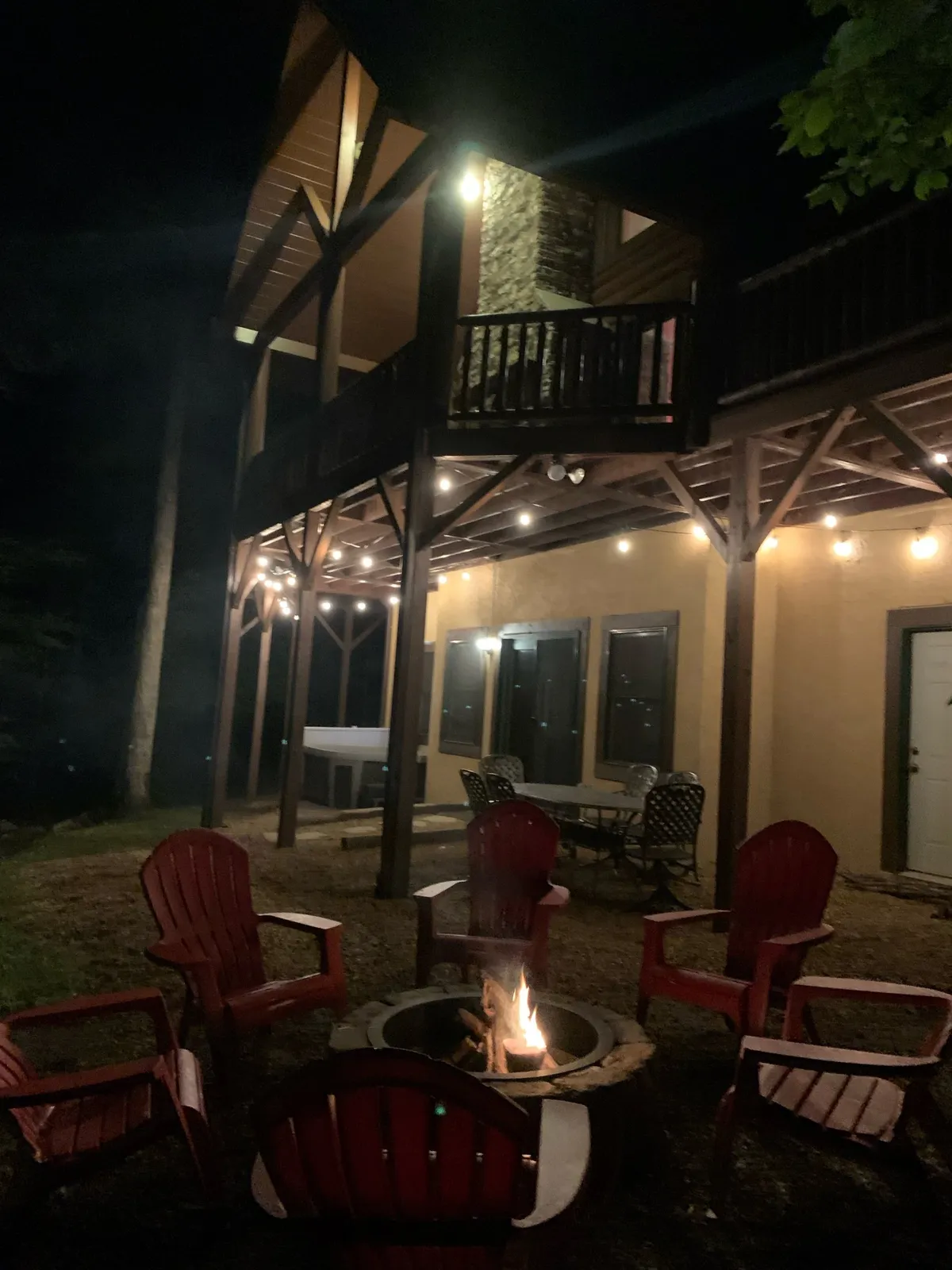 An inviting exterior space with a fire pit encircled by vibrant red Adirondack chairs, string lights hanging above, and the house in the background. The setting is designed for group gatherings, evening relaxation, and outdoor enjoyment.