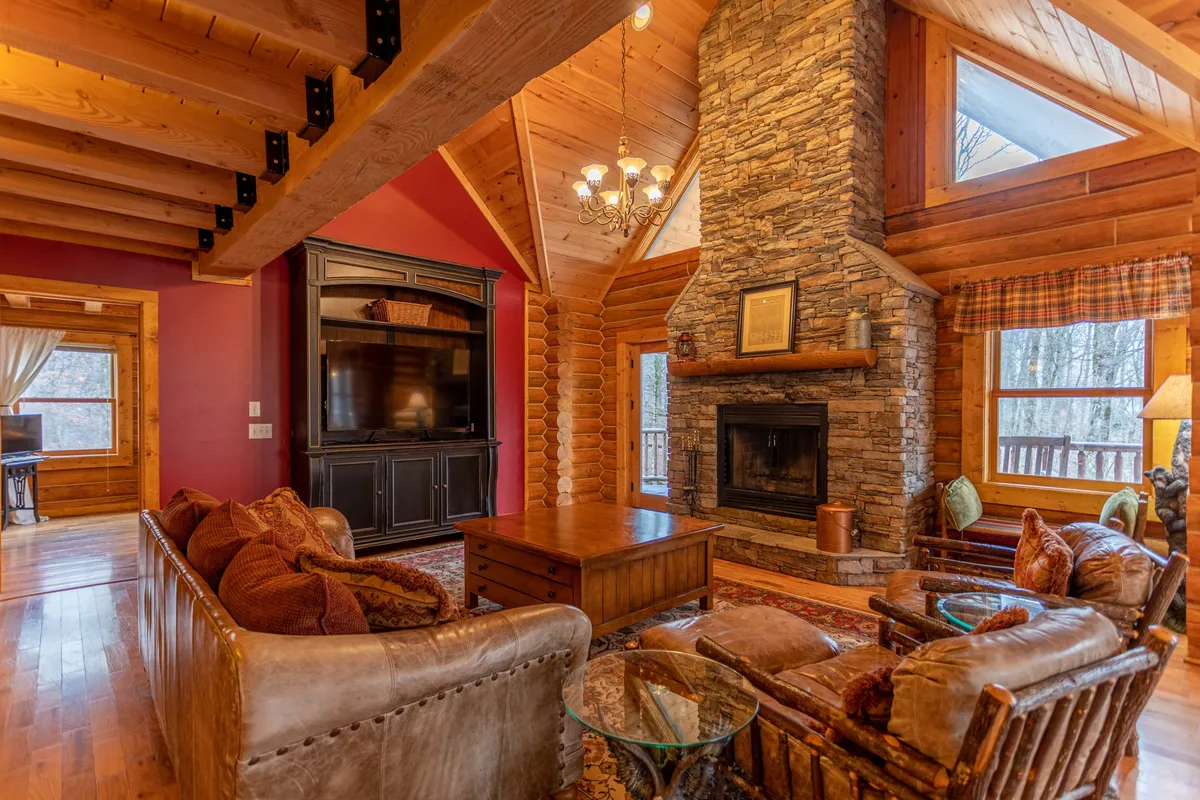 A spacious living room featuring a majestic stacked-stone fireplace, exposed wooden beams across vaulted ceilings, plush sofas arranged for comfort, and a large flat screen TV set within a warm, inviting ambiance. Natural light streams through expansive windows, emphasizing the cozy yet grand atmosphere perfect for group gatherings.