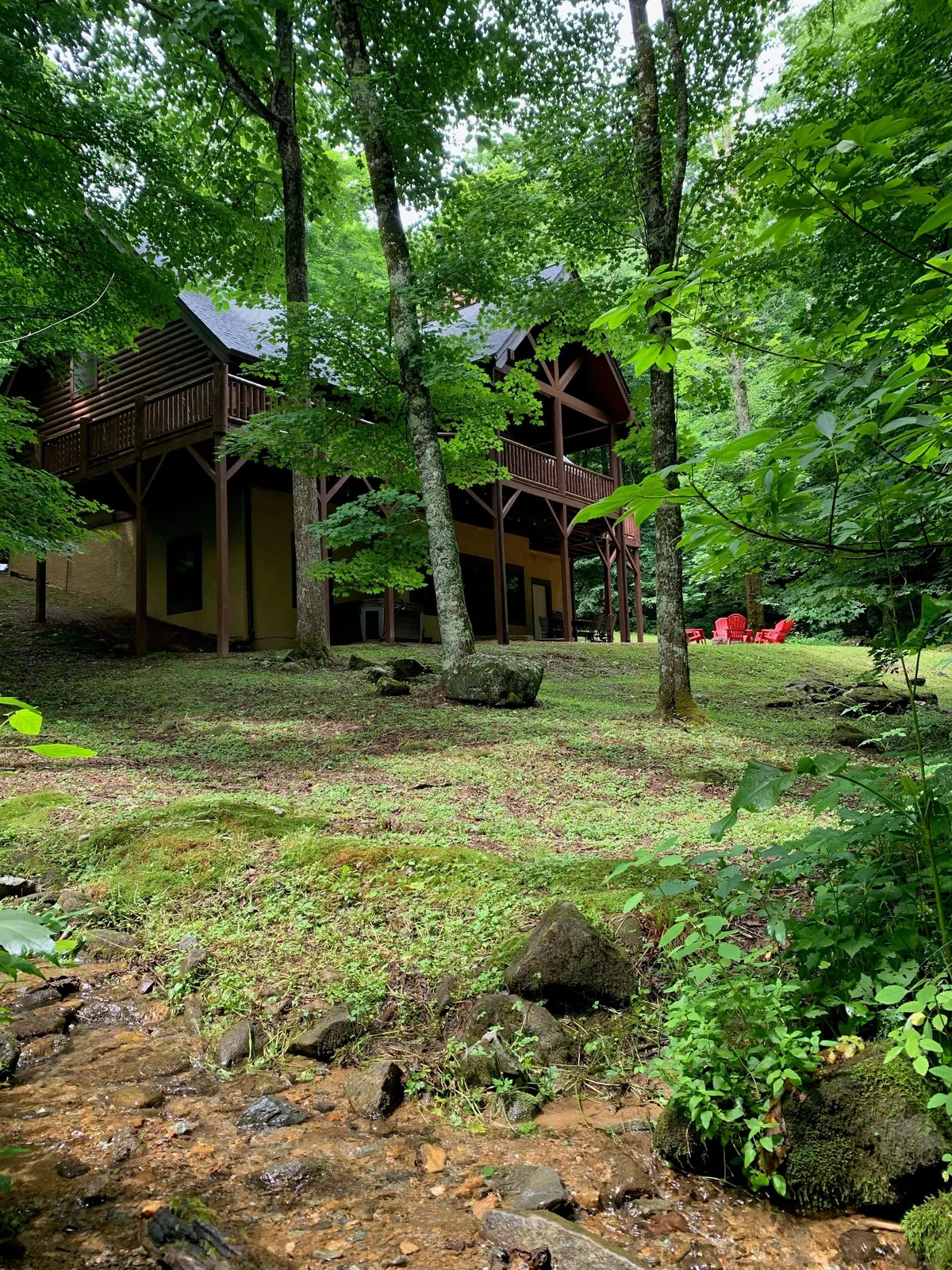 A picturesque exterior view of the wooden house surrounded by lush greenery, featuring a stream in the foreground and inviting red chairs set out for relaxation. The scene conveys a peaceful outdoor environment ideal for both people and their pets to explore or unwind by the water.